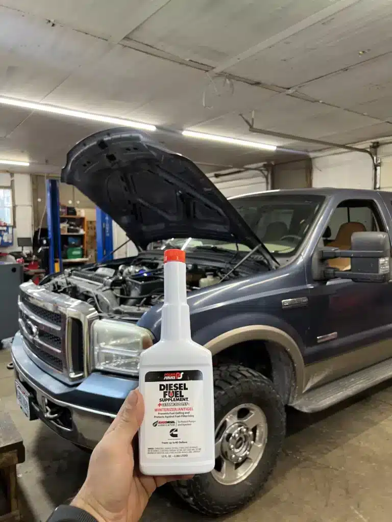 A person holds a bottle of Power Service Diesel Fuel Supplement in front of a dark-colored pickup truck with its hood open inside a garage or workshop.