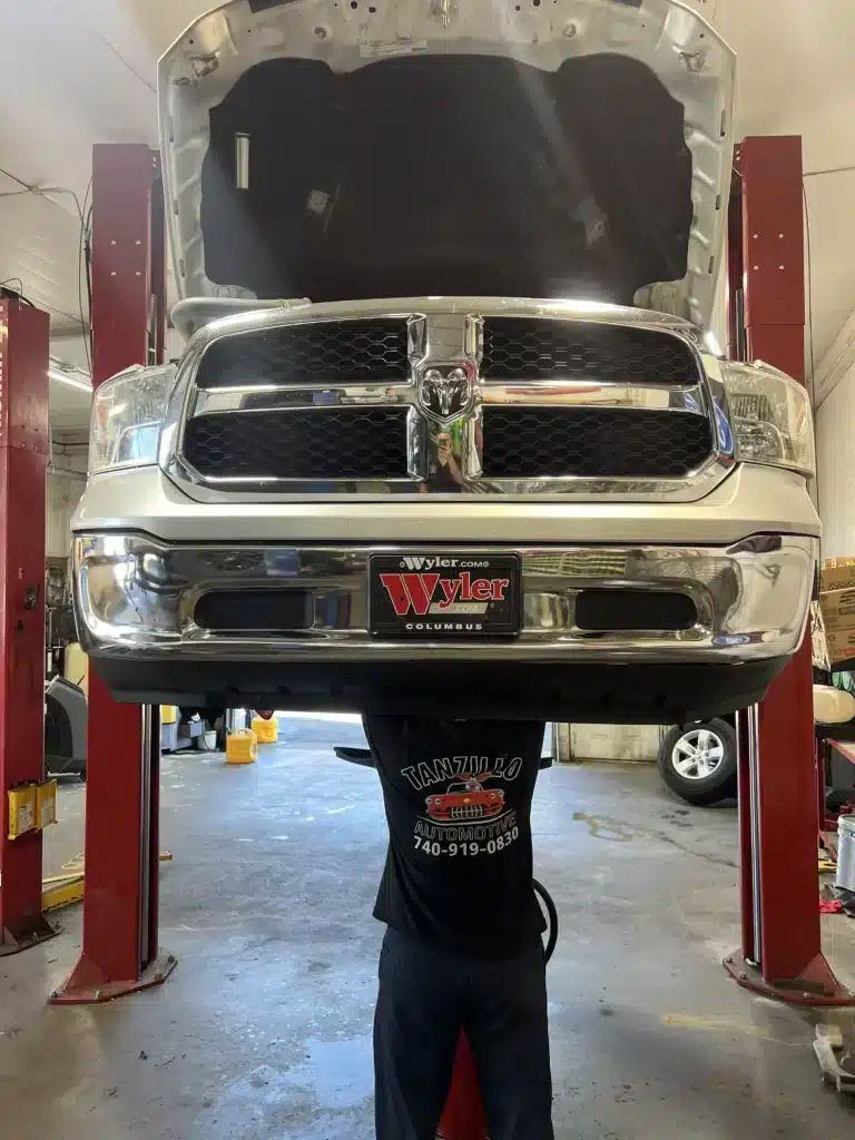 A mechanic appears to hold up the front of a white Dodge Ram truck raised on a lift in a garage, with the trucks hood open and a Wyler Columbus license plate visible.