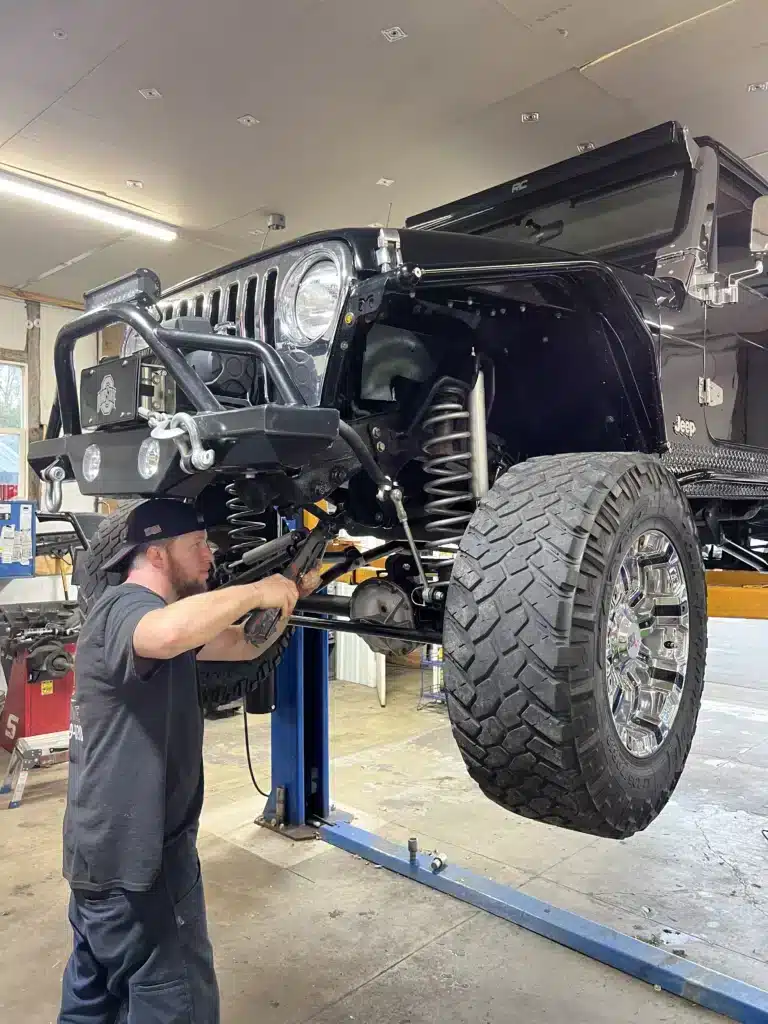 A mechanic wearing a black hat works on the raised front end of a black Jeep with large off-road tires and a lifted suspension inside a garage. The Jeep is elevated on a blue hydraulic lift.