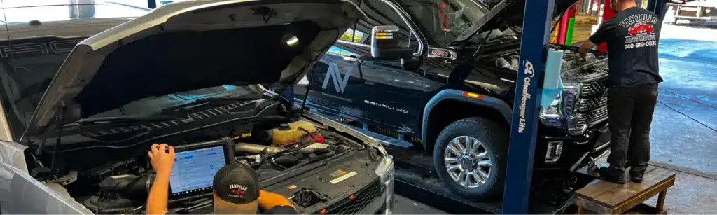 Two mechanics work in a garage; one uses a laptop to diagnose a white vehicle with its hood open, while the other inspects a raised black truck. Tools and equipment are visible in the workshop.