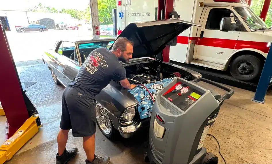 A mechanic in shorts and a t-shirt works under the hood of a classic car hooked up to an AC service machine inside a garage; an ambulance is parked beside the car.