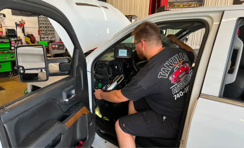 A mechanic in a black T-shirt works inside a white vehicle with the driver’s door open and the hood raised in an auto repair shop, using diagnostic tools on the dashboard.