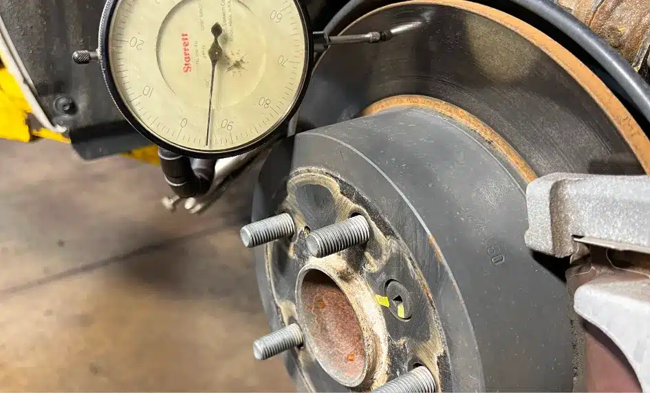 A dial indicator measures the runout of a car brake rotor, with the gauge tip touching the rotor surface near the wheel studs on a vehicle in a garage setting.