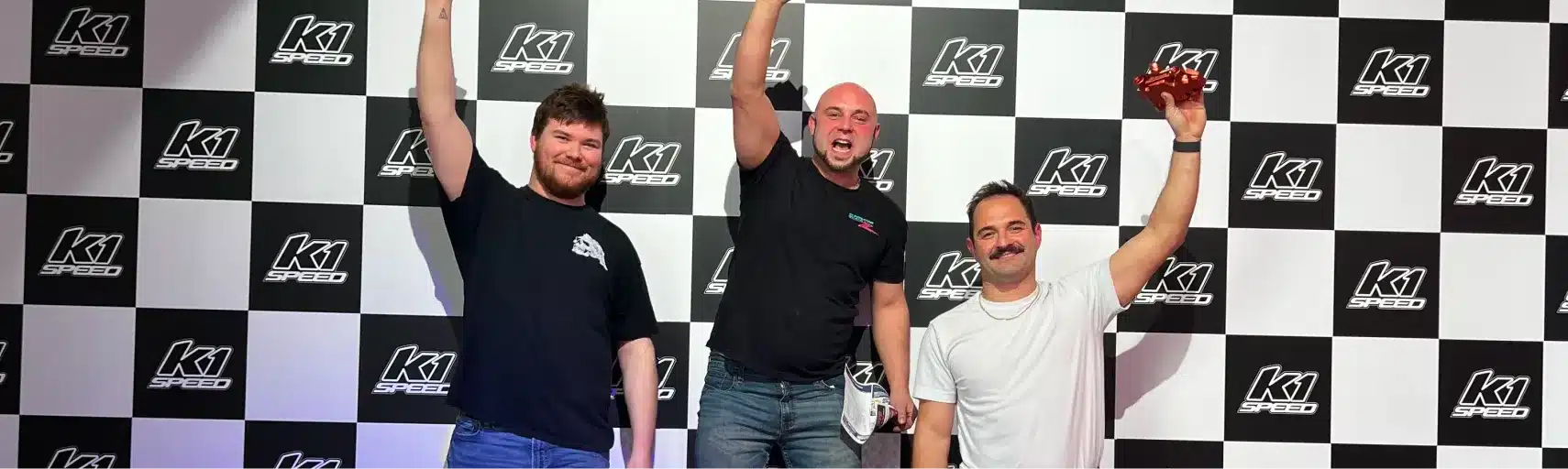 Three men stand on a winners podium in front of a K1 Speed checkered backdrop, smiling and raising one arm each in celebration. The man in the center is in first place, flanked by second and third place winners.