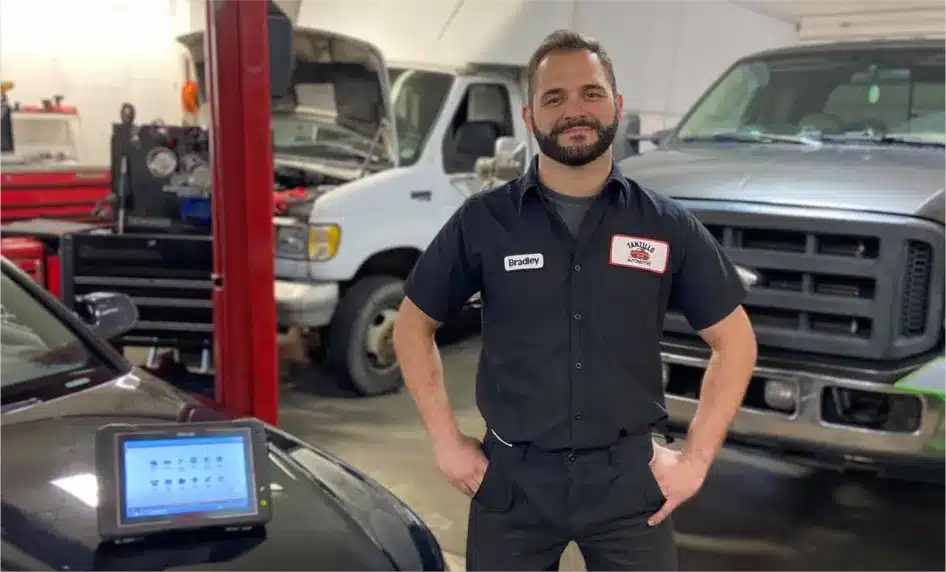 A mechanic wearing a black uniform with a name tag stands smiling in an auto repair shop, surrounded by vehicles and equipment. A diagnostic device sits on the hood of a nearby car.