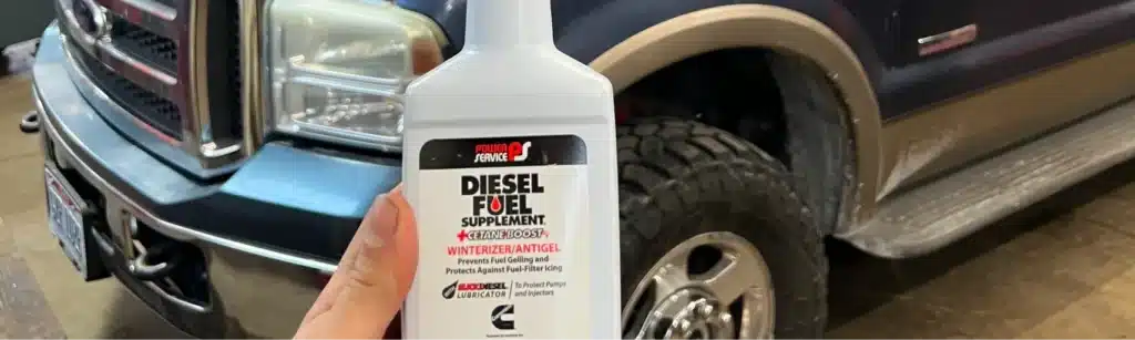 A hand holds a bottle of Power Service Diesel Fuel Supplement in front of a blue pickup truck with large tires parked in a garage. The bottle label mentions anti-gel and winterizer features.