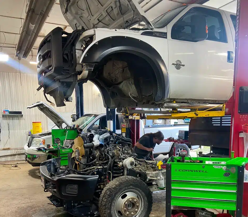 A mechanic works on the exposed engine and frame of a white pickup truck lifted on a hydraulic car lift in an auto repair shop, with tools and equipment nearby.