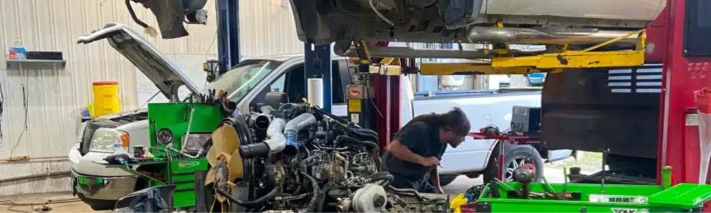 A mechanic works on a car engine in a busy auto repair shop, with vehicle parts, tools, and equipment scattered around. Multiple vehicles are lifted and being serviced in the background.