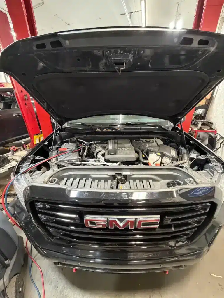 Open hood of a black GMC truck in a garage, revealing the engine and various components. Red support pillars and tools are visible, suggesting maintenance work.