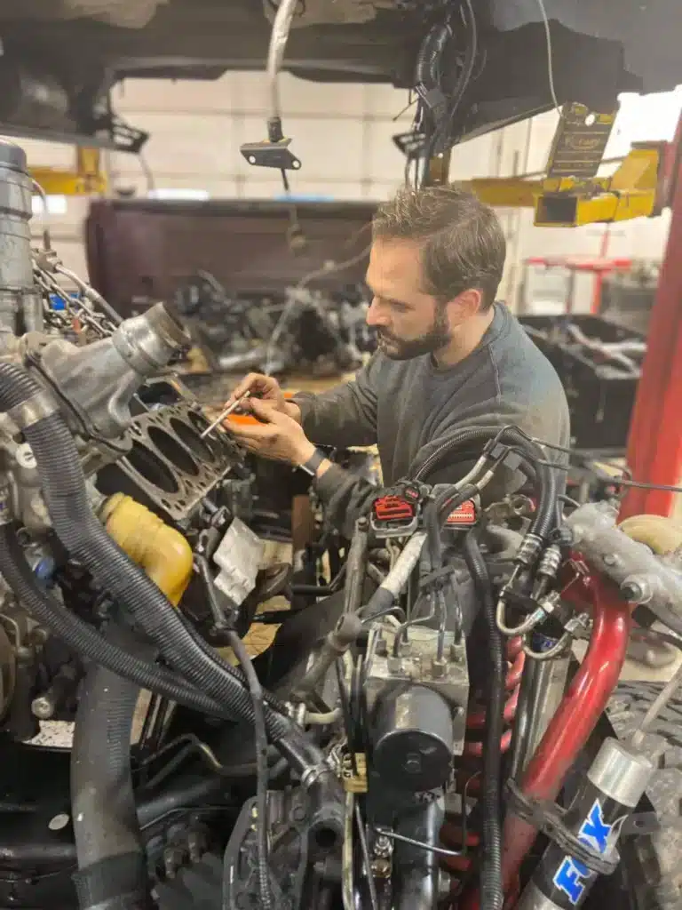 A mechanic with a beard focuses intently on repairing a vehicle engine in a workshop. The scene conveys concentration and technical skill amidst various mechanical parts.