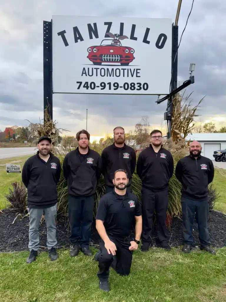 Six men in black shirts stand in front of a Tanzillo Automotive sign outdoors. Overcast sky, autumn foliage; conveys a friendly team environment.