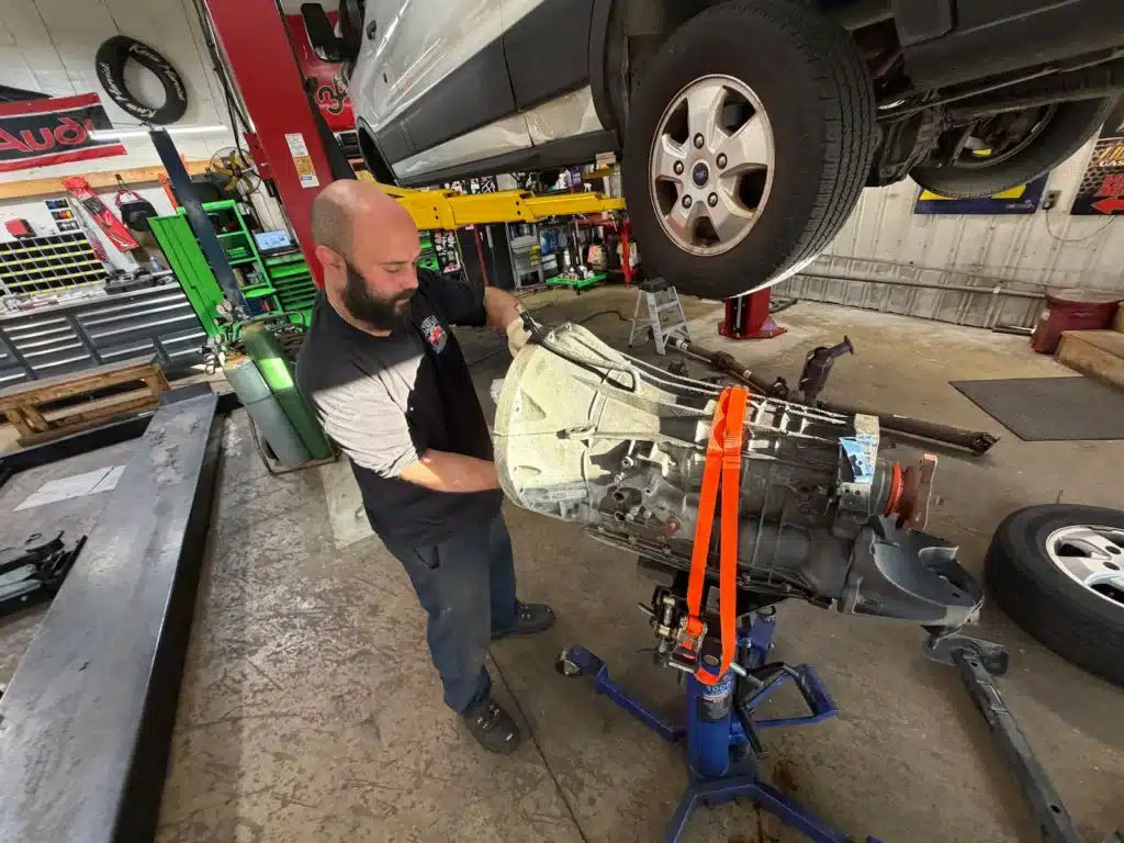 A mechanic in a garage works on a lifted car's transmission using a hoist. The setting is well-equipped, conveying focus and expertise.