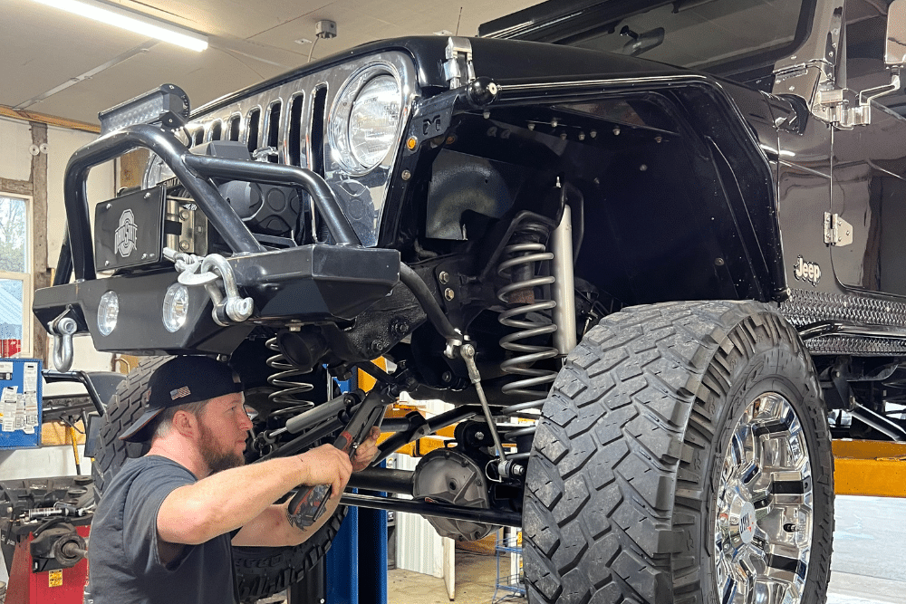 Steering and Suspension, Auto repair in Pataskala, OH. Mechanic working under a lifted Jeep in a repair shop, inspecting suspension and electrical components that can affect battery performance.