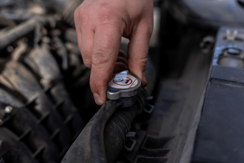 Cooling System Service in Pataskala OH At Tanzillo Automotive. A hand is gripping a radiator cap under the hood of a vehicle, preparing for a cooling service repair. The engine components are visible, and the cap has a warning symbol indicating not to open when hot—a crucial safety reminder for anyone performing Pataskala cooling system service.